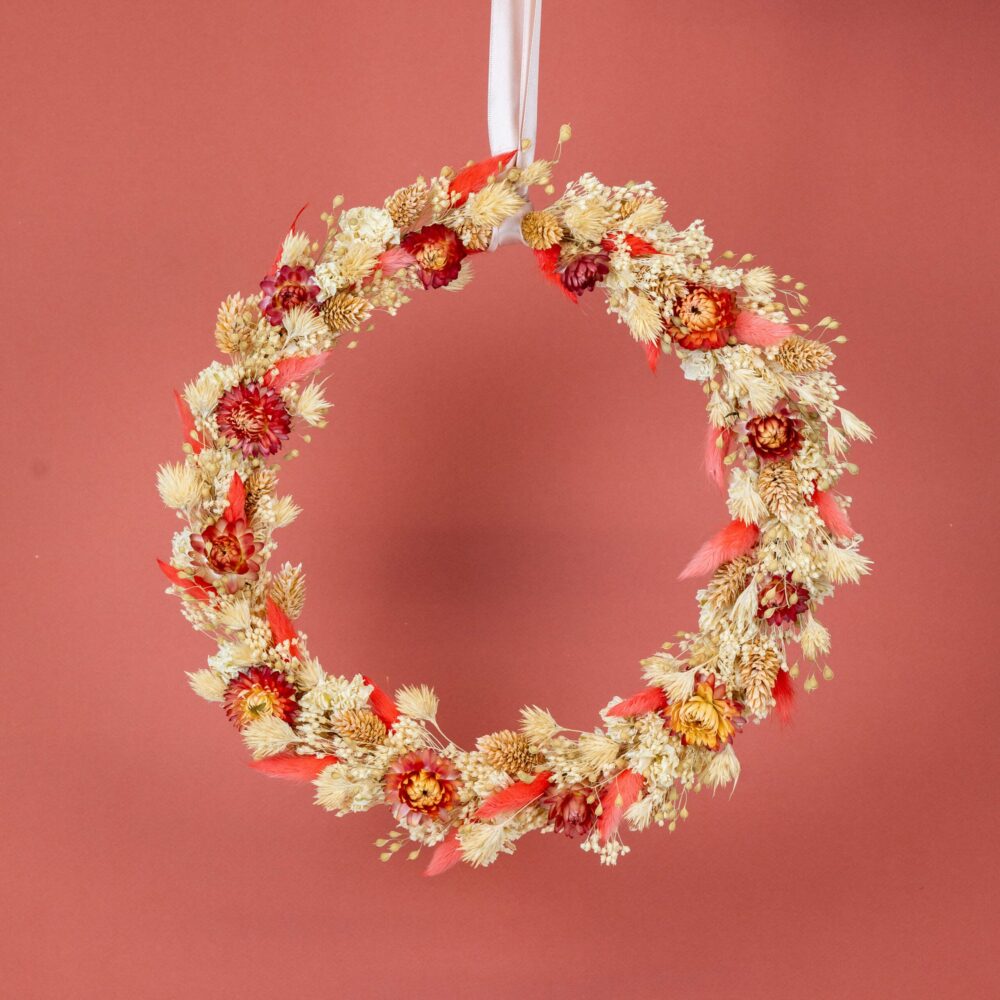 une couronne de fleurs séchées suspendue par un ruban blanc. La couronne est ornée de petites fleurs rouges et jaunes, d'épis de blé doré et de plumes rouges, le tout arrangé sur un fond rose terre cuite. Les textures et les couleurs naturelles créent une ambiance chaleureuse et festive.